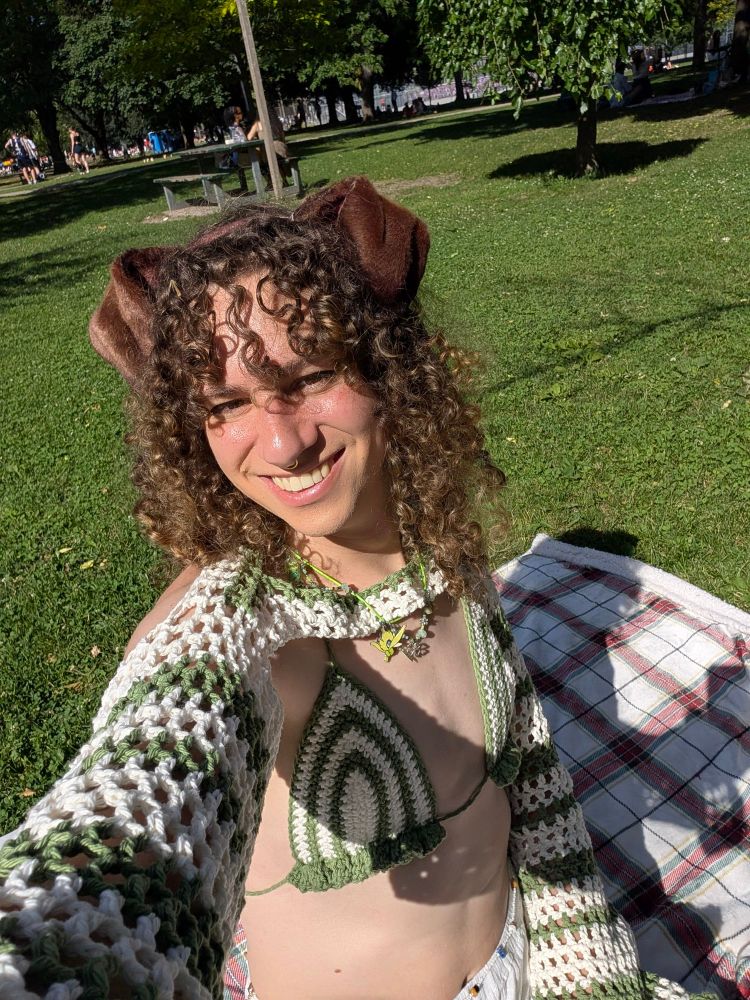 a selfie photo of ari. she's in a park, there's trees and people in the bathroom, and grass. there's a blanket on the ground. she's wearing a green and white crochet bikini top & sleeves, and she's got dog ears. she's smiling. 