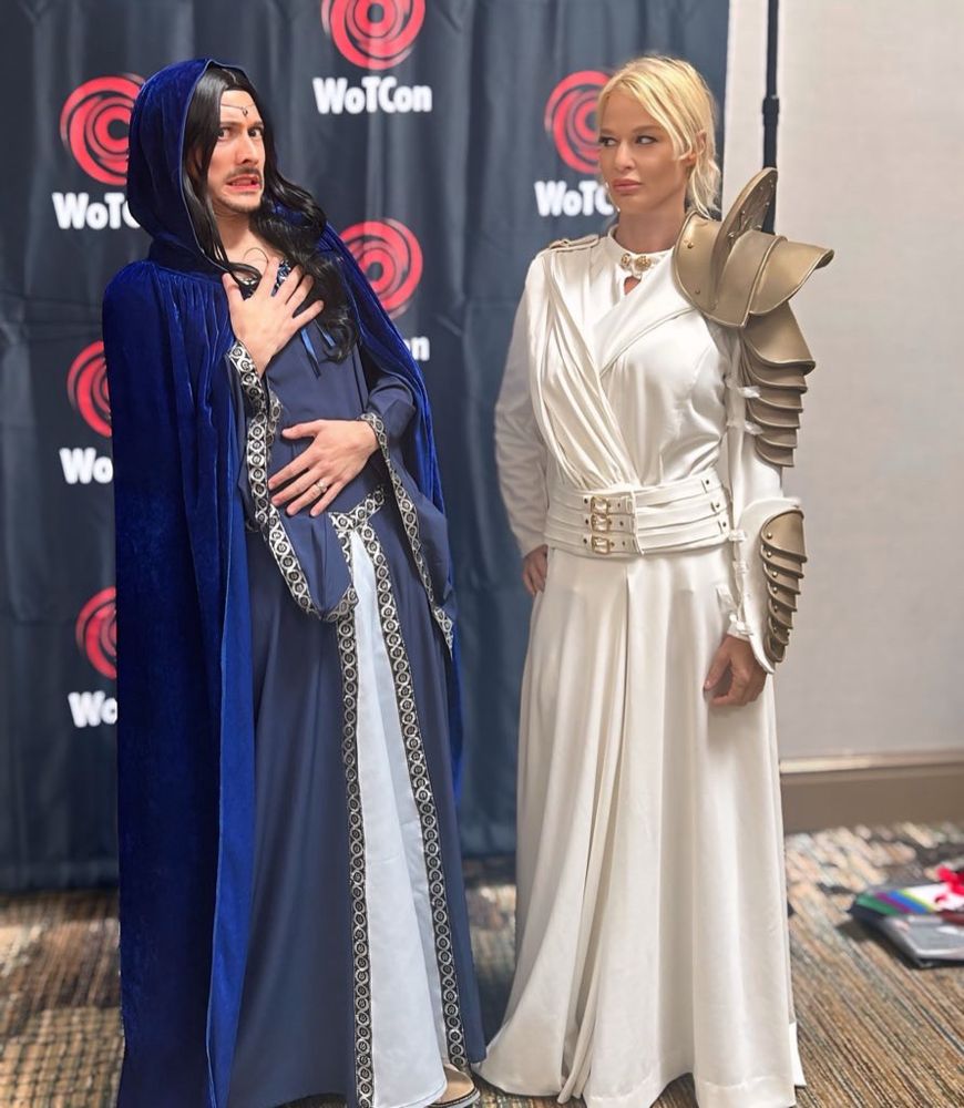 Moiraine in blue medieval style gown with blue cloak and a blue headband with a stone and a mustache with London in white whitecloak cosplay and gold armor on the left arm