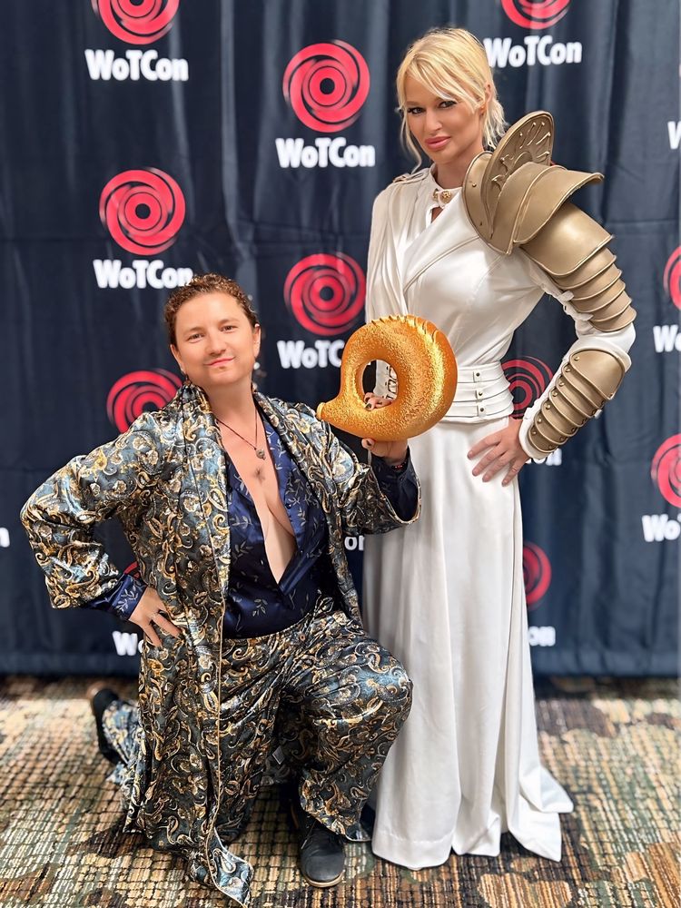 Dovie as Mat in blue and gold satin pajamas, a mustache, a tuft of chest hair, kneeling with horn of valere and London as a whitecloak from season one with gold armor on left arm 