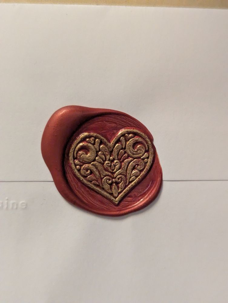 A red wax seal on a white envelope. The seal is in the shape of a heart. The heart is covered in gold leafing marker, making it gold colored to stand out from the red wax.