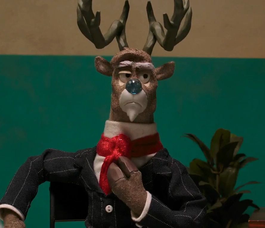 An anthropomorphic reindeer with a blue nose, wearing a black suit, white button up shirt beneath it, and a red tie.