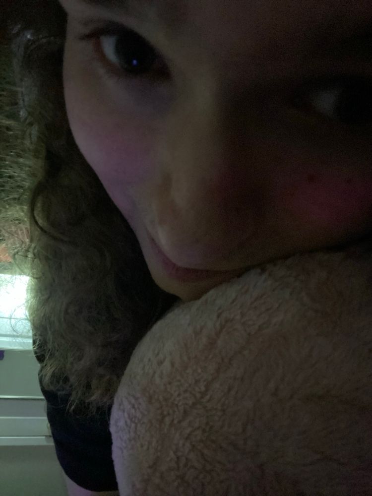 A close up selfie of a white woman snuggling a pink shark plush. She is smiling and her eyes look huge as they look directly at the viewer.