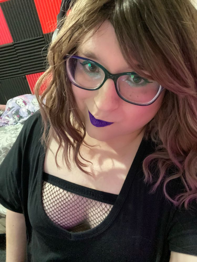 A picture of a white woman with shoulder length brown hair, wearing a low cut black top with fishnets underneath showing her massive cleavage. She is smiling at the camera with lips colored with purple lipstick, dark green makeup around her eyes.