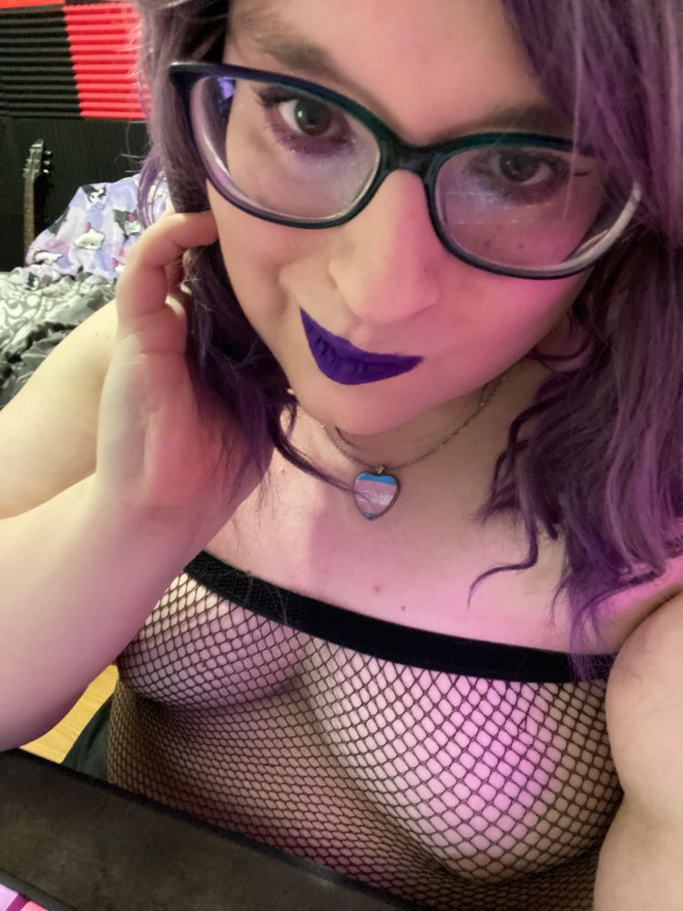 a white woman wearing nothing but fishnet pulled up above her breasts is looking seductively at the camera with one hand in her shoulder length hair. 