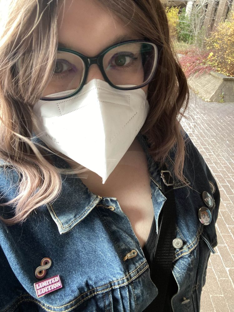 A photo of a white woman with shoulder length brown hair standing on a brick walkway, with some foliage behind her. She has big rimmed glasses and purple eyeshadow. She is wearing a white KN95 mask