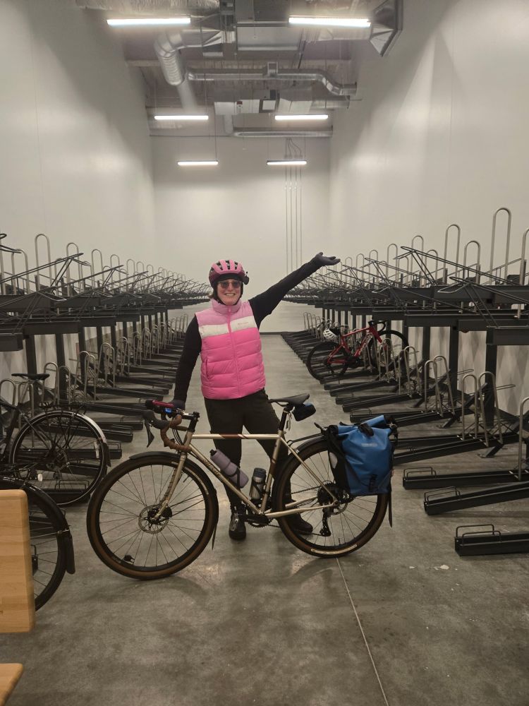 Woman in pink puffy vest and pink bicycle helmet poses behind bicycle inside bike room