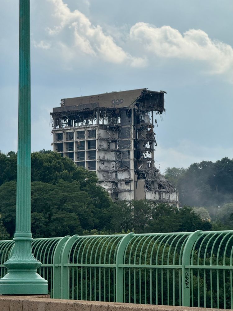 The last of a crumbling former hotel—the Key Bridge Marriott, closed in 2021 and condemned in 2023. Still 12 stories tall but now only a couple rooms deep… and you might not enjoy the amenities.

Between the hotel and the camera are leafy green trees. In the foreground is the teal railing, and one of the light-posts, of Key Bridge itself.