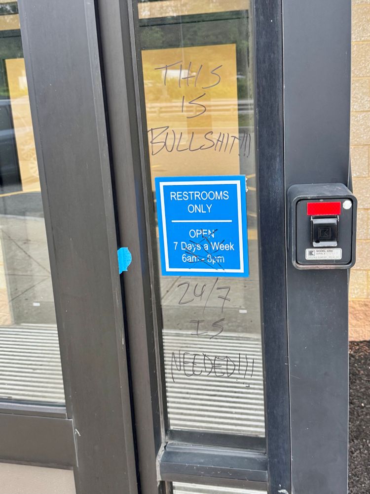 On a highway rest stop, next to the door, a sign reads “RESTROOMS ONLY - OPEN 7 Days a Week 6 a m to 8 pm.” The open hours are scribbled over with black marker. In the same black marker, above and below the sign, is written “This is bullshit! 24/7 is needed!!!”