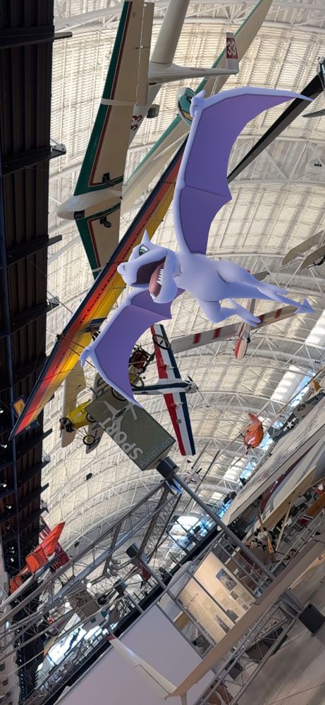 In the massive hangar of the National Air & Space Museum’s Udvar-Hazy Center, hang gliders and other small sport aircraft are on display, suspended in the air. Soaring alongside them is Aerodactyl, a grey pterosaur Pokémon, with its wings spread in parallel to the nearest hang glider.