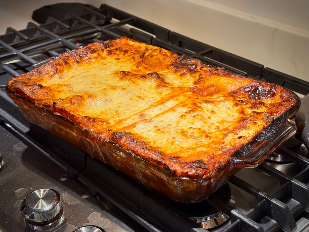 A gorgeous golden-brown lasagna bubbles just above the edge of a casserole dish, fresh out of the oven, sitting on the stovetop to cool.