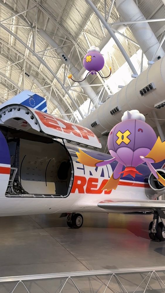 Drifloon and Drifblim, purple-red-white-and-yellow balloon Pokémon, float happily in the air in the huge hangar that is the National Air & Space Museum’s Udvar-Hazy Center. Behind them is a purple-red-and-white Federal Express jet airplane on display.