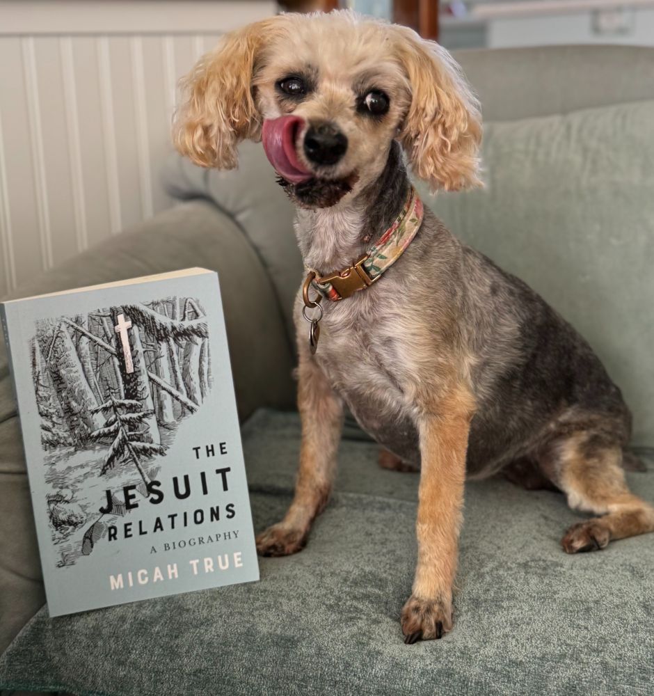 small dog licking its chops next to a book "The Jesuit Relations a Biography" by Micah True