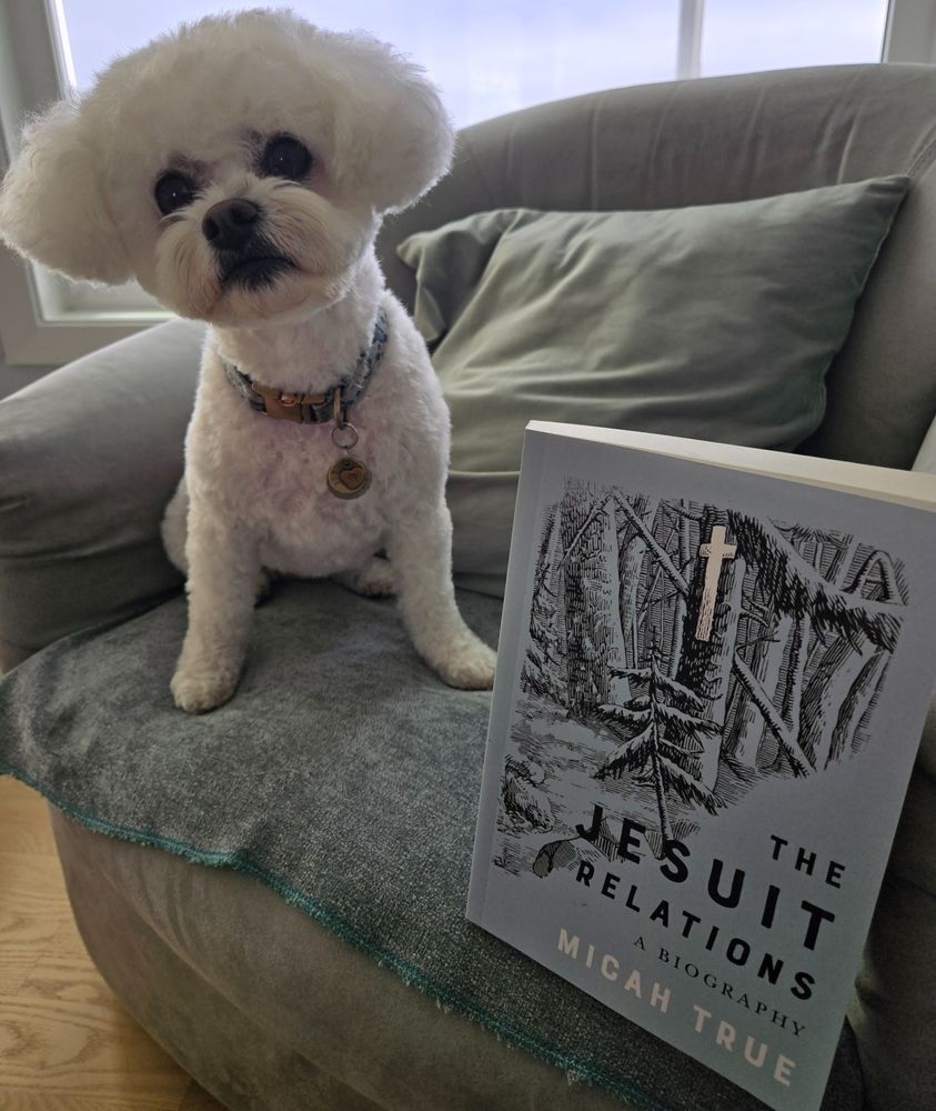 small white bichon frisée sitting on a chair next to a book entitled "The Jesuit Relations a Biography" by Micah True