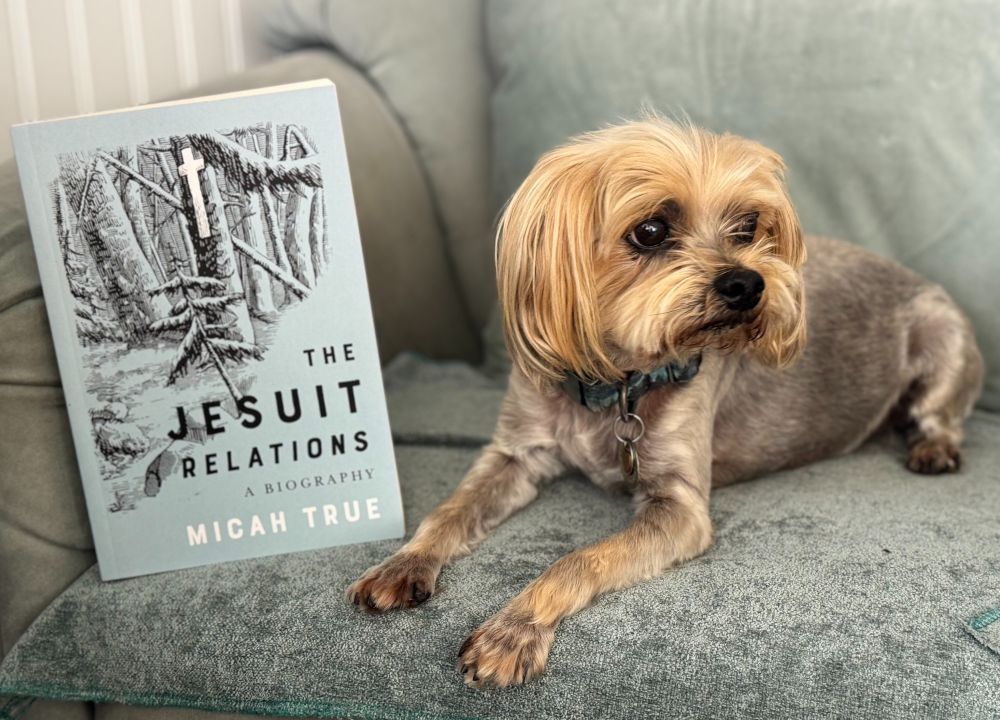 small dog lying on a chair next to a book entitled "The Jesuit Relations a Biography" by Micah True