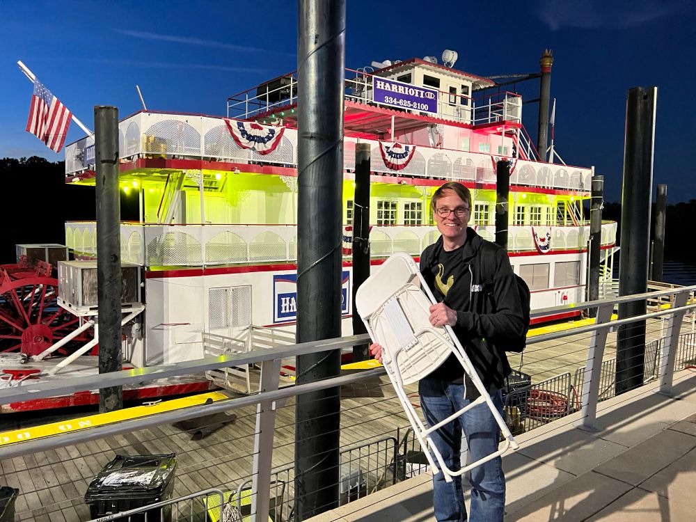 A guy in a Spurs shirt holds a white folding chair on a dock in front of a riverboat.