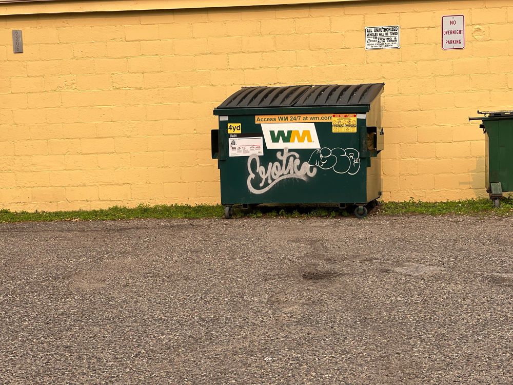 A green dumpster with “Erotica” graffitied on it in gray script sits in front of a yellow wall.