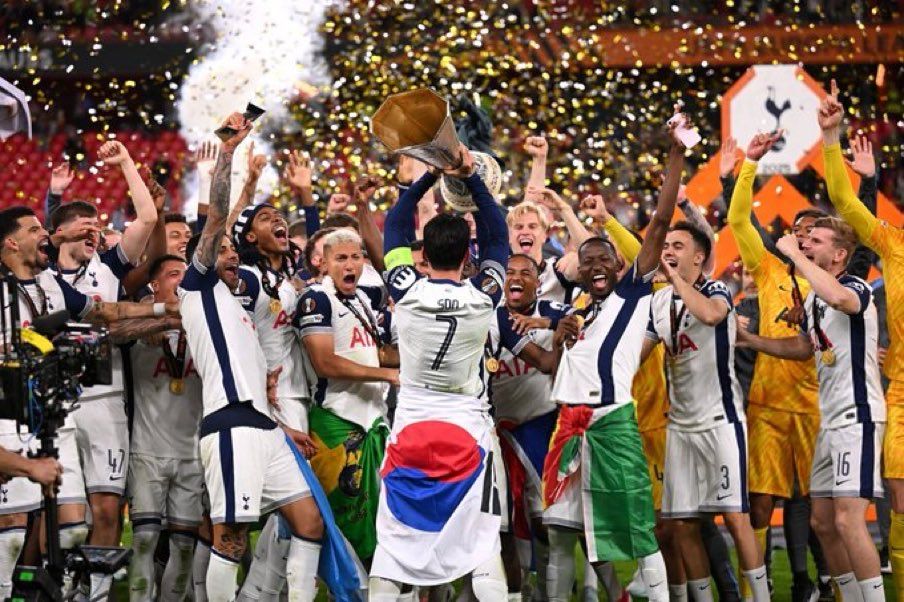 Son Heung-min wrapped in a South Korean flag lifts the Europa League Champions Trophy in front of the rest of the celebratory Tottenham Hotspur team. Confetti flies in the background.