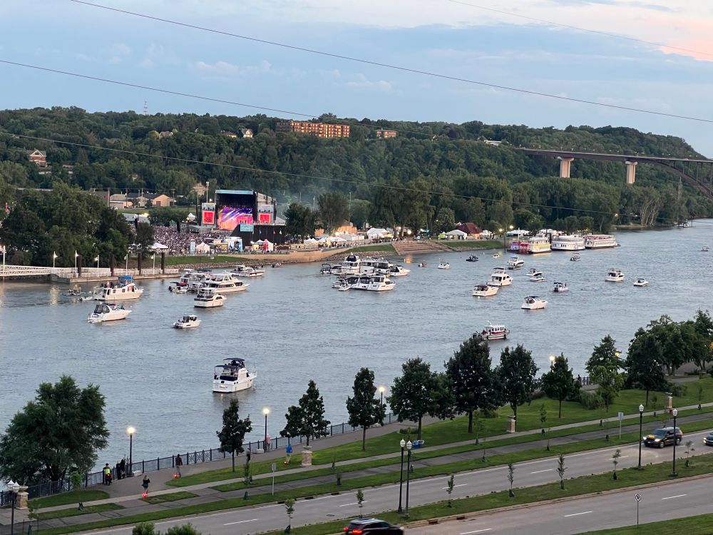 The Mississippi River in St. Paul with a lot of boats. On the other side of the river a large stage with Fall Out Boy performing has many people in front of it.