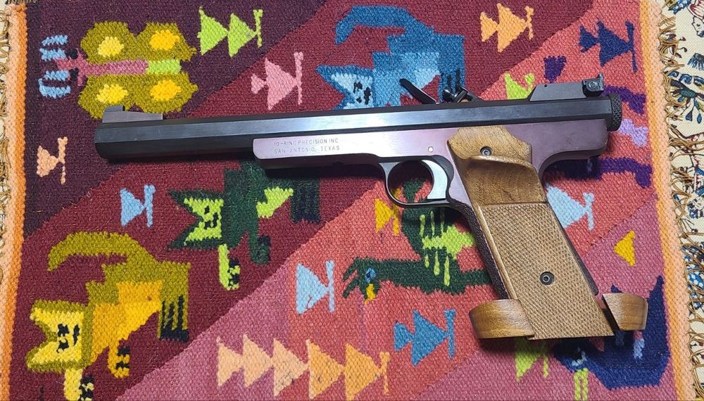 A left side photograph of an unusual target pistol. The design is strikingly modern, similar to single-shot Olympic pistols, but the operating mechanism is a flintlock, a firearms action typically considered obsolete, used in antique replicas.
The light color wooden grips are highly contoured, with a prominent thumb rest and palm shelf.
The barrel is black, octagonal in diameter, and strongly rifled. The frame is solid steel with a burgundy finish.

