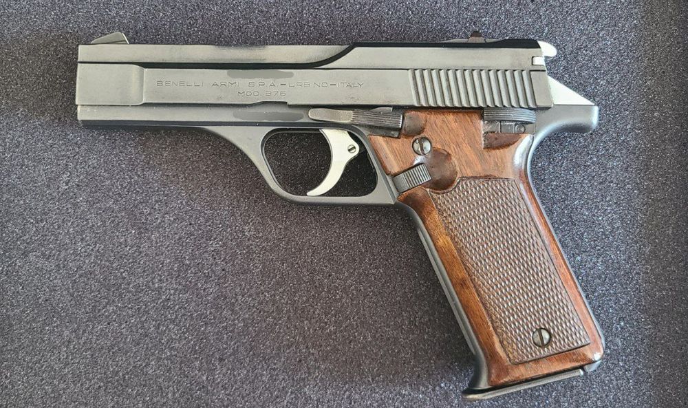 A left side photograph of a handgun. The slide is quite rectangular, with bold lines and angles. This side of the pistol has the controls. The safety, slide release, and magazine release each have their own scalloped surfaces in the wood grips. 
The serial numbers on the frame and slide have been digitally removed in the image. 