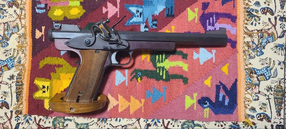 A right facing photograph of the same unusual target pistol. The flintlock action is  on this side and it is on the exterior of the pistol, directly above the trigger.  

The frame is very similar to a 1911A1, with existing scalloped cuts near the trigger, but otherwise solid. Also, the trigger pivots instead of sliding. Single action is somewhat heavy but trigger travel is barely measurable in millimeters.