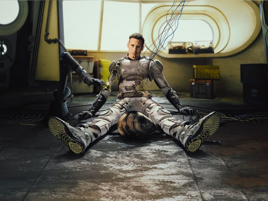 Murderbot from Apple TV sitting on the floor in science fiction habitat. It has significant damage and wires hanging out