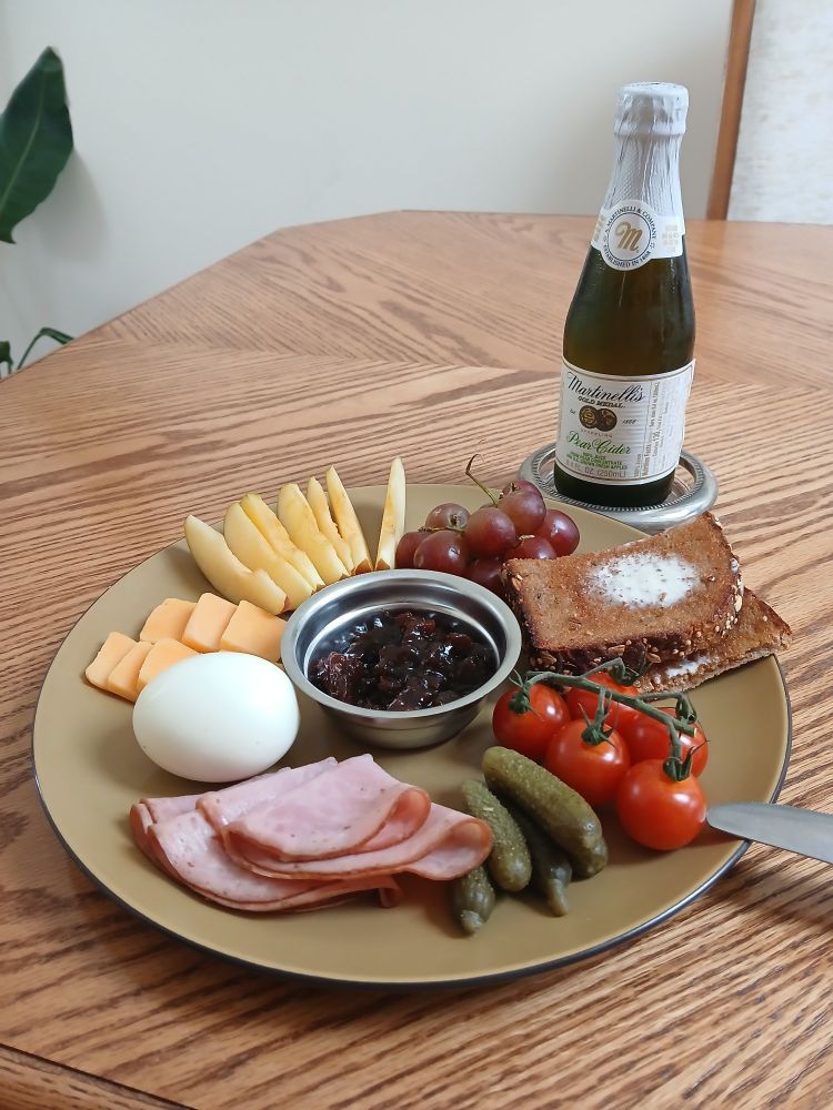 A ploughman's lunch on a large olive-green plate. Clockwise: red grapes, toasted and buttered whole grain bread, cherry tomatoes on the vine, cornichons, honey smoked ham, a hard-boiled egg, a few small slices of cheddar, and some apple slices, all surrounding a small dish of Branston pickle. There is also a small bottle of Martinelli's pear cider.