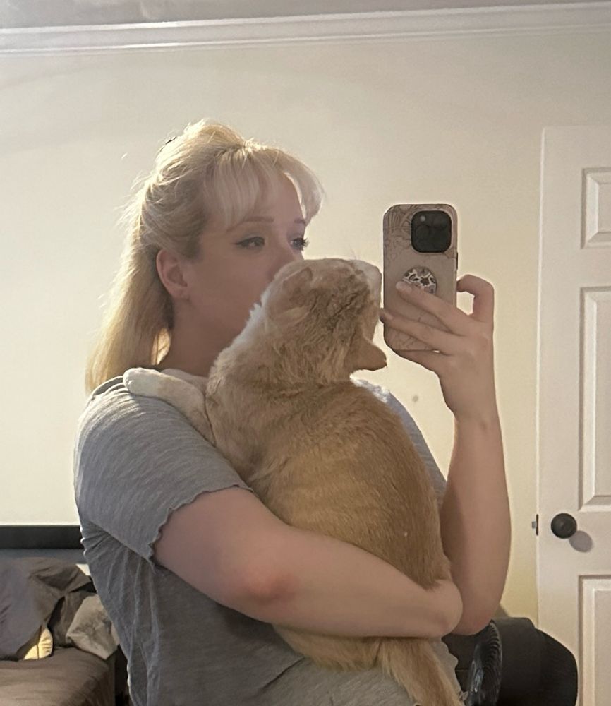 me, a blonde white woman, holding my orange cat while taking a picture in the mirror 