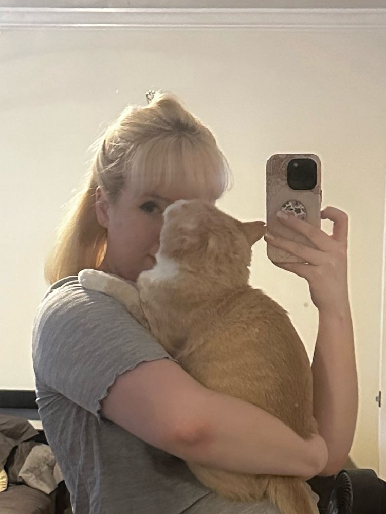 me, a blonde white woman, holding my orange cat while taking a picture in the mirror 