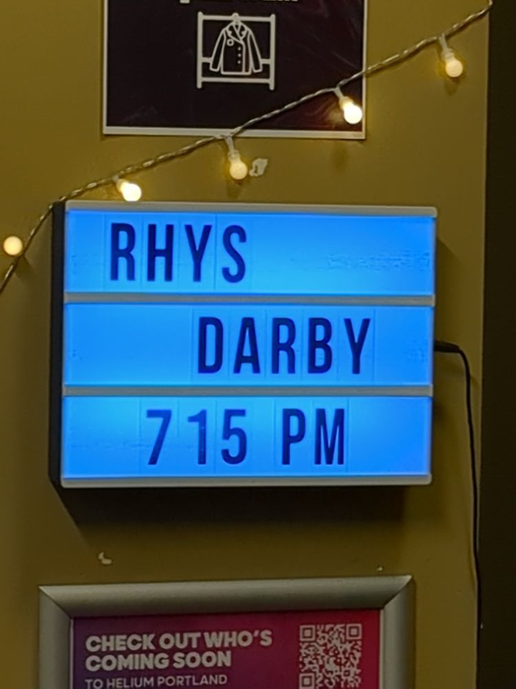 One of those light up mini marquis signs on a yellow wall, the letters spell out RHYS DARBY 7:15 PM. 