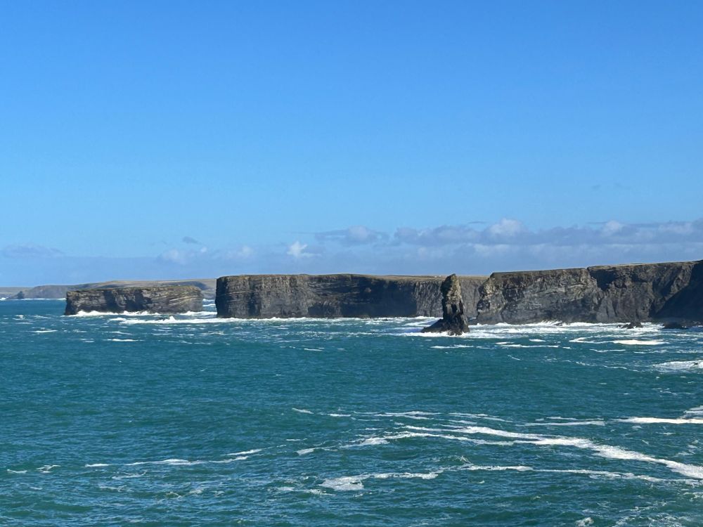 Cliffs of Kilkee, west coast of Ireland. Gray cliffs rise dramatically from a blue ocean against a blue sky with a few purple clouds with a weathered sea stack just offshore.