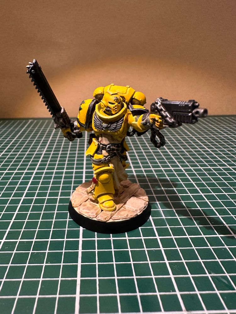 A space marine in yellow armour aims a bolter in with hand and carries a chain sword in the other. 