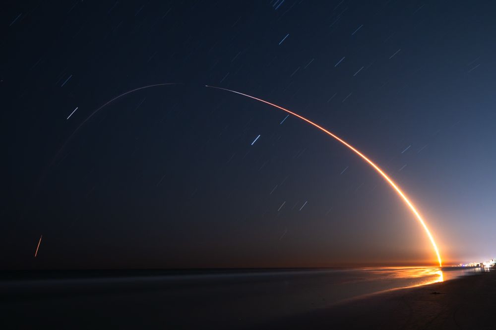 The light streak of a rocket launch arcs over a beach, illuminating the water. Faint, short star trails are visible in the sky. 