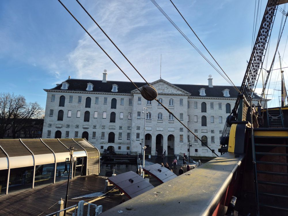 Het scheepvaartmuseum in Amsterdam gezien vanaf de replica van VOC schip de Amsterdam. Op de achtergrond het museum onder een mooie winterblauwe lucht, op de voorgrond de boordrand en staand want van de Amsterdam.