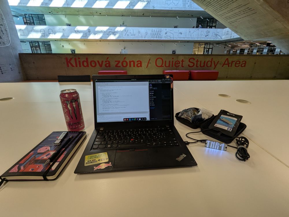 Photo of a desk in a public library. There is a laptop, a journal, a can of energy drink and case with small electronic parts on the desk. The laptop is running IDE and there is a LED matrix connected to it