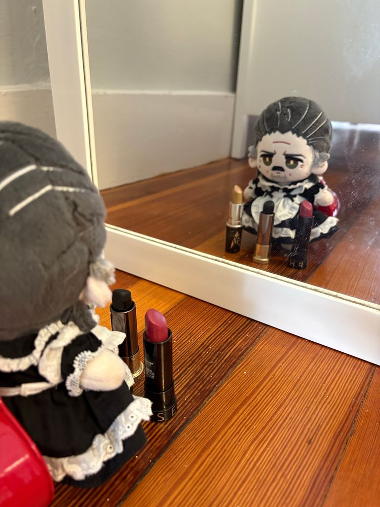 A 15cm Izzy doll by Rights in a black maid’s uniform looking in a mirror with gold, black, and pink lipsticks in front of him. 