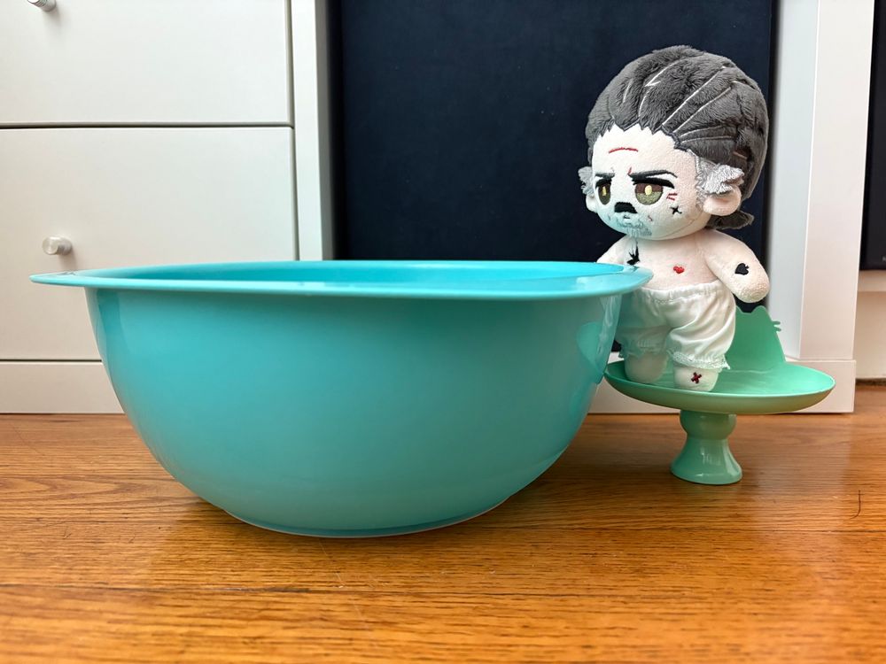 A 10cm Izzy dolls by Rights wearing white bloomers stands on top of a pusheen candle holder next to a large blue mixing bowl, he looks like he’s looking in the water. It’s on a brown wooden floor with shelving units behind it. 