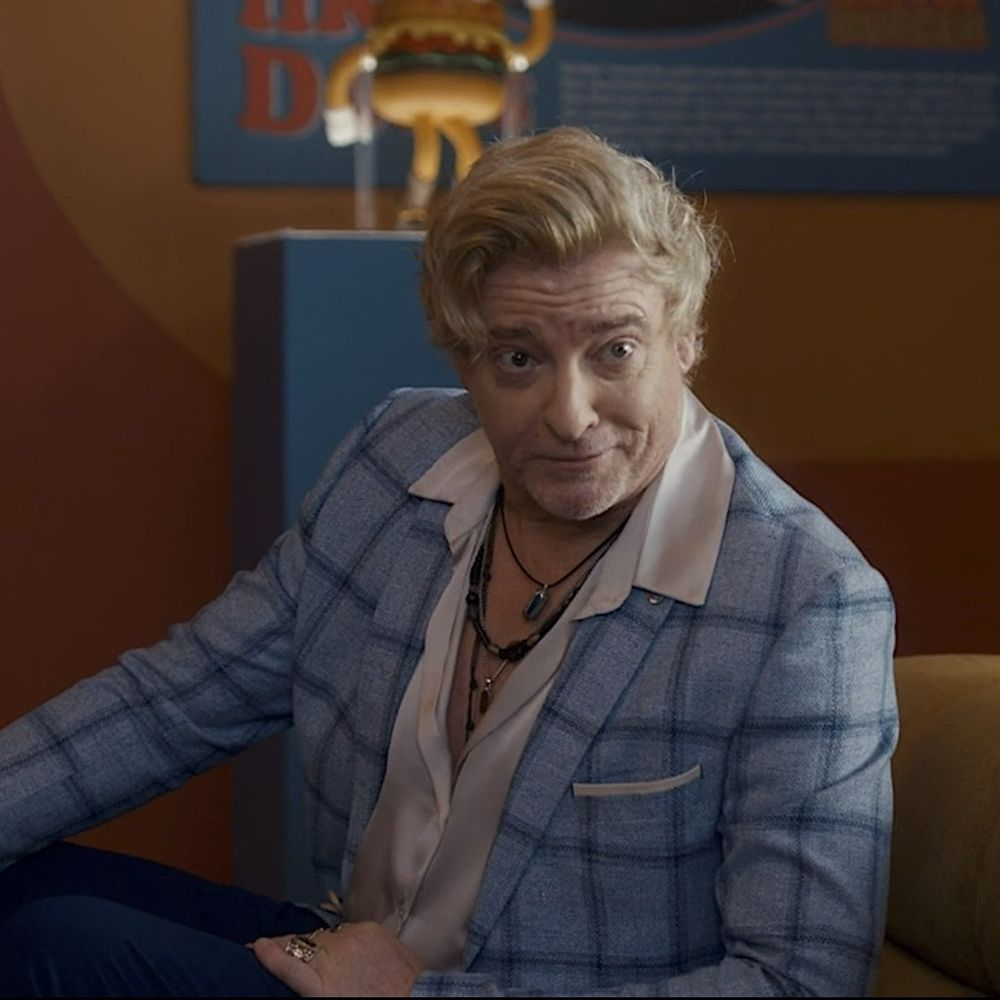 Rhys Darby in a blue suit, with unbuttoned shirt. Rhys is leaning forward with a smirk 