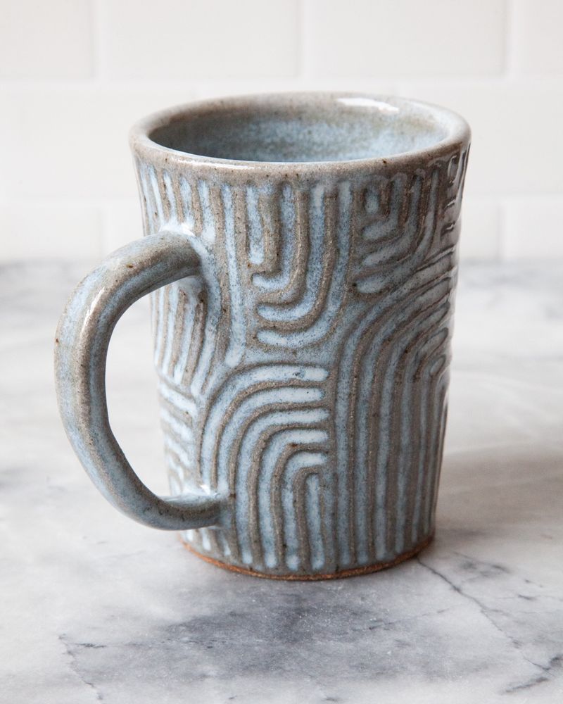 A light blue carved ceramic mug. The mug is sitting on a white marble surface with a white tiled background.