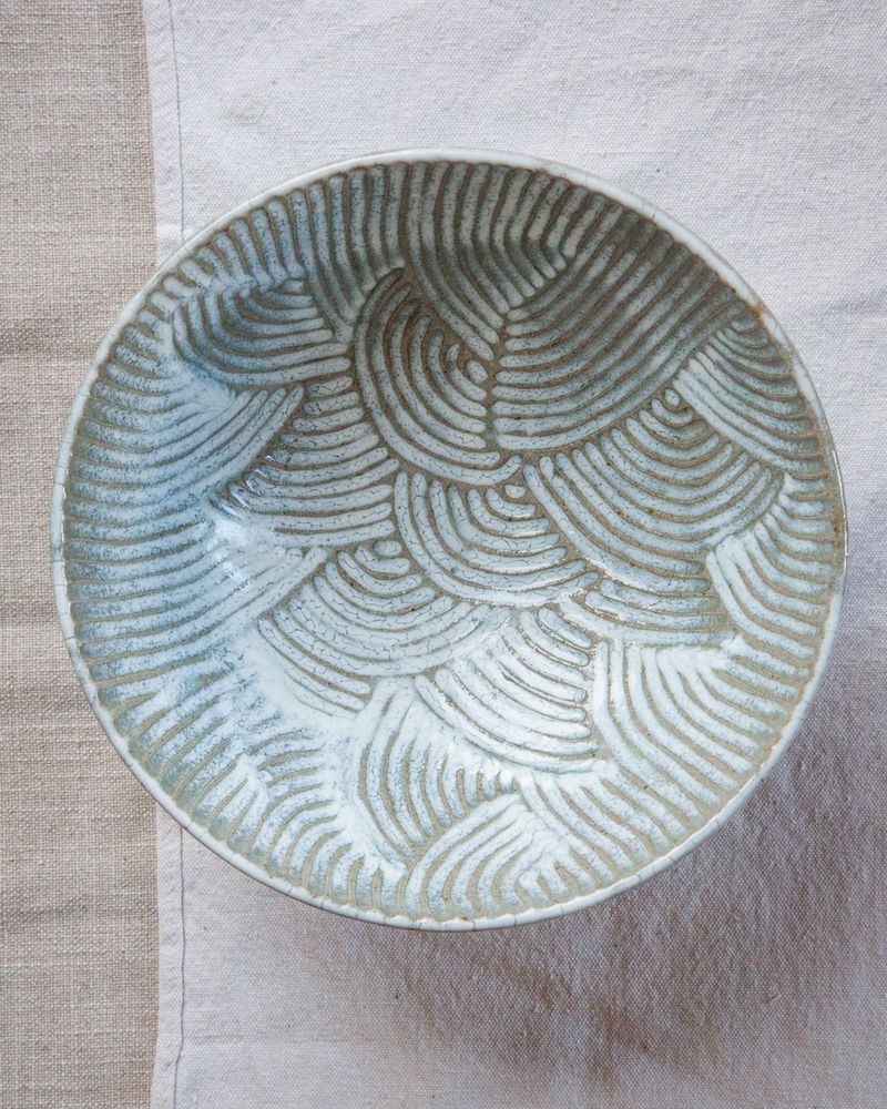 A light blue and white carved large ceramic bowl sitting on two pieces of beige fabric.