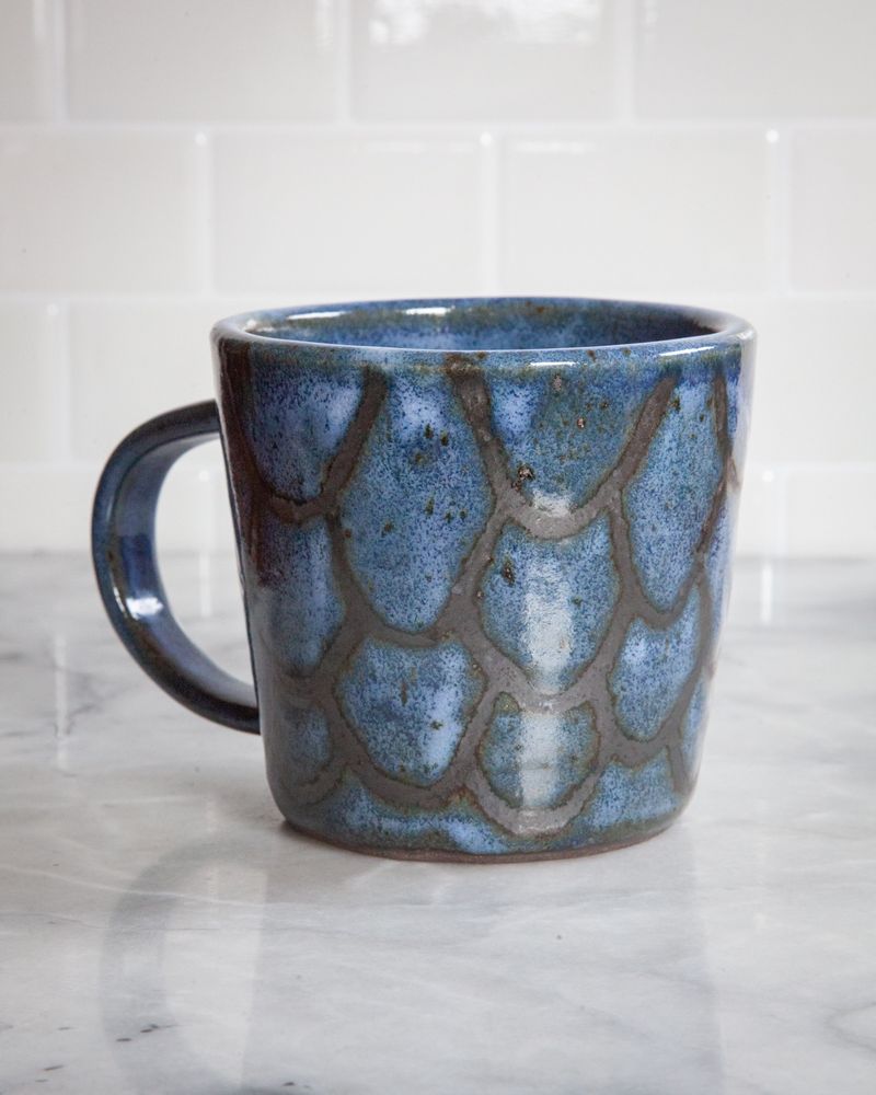A blue ceramic mug with black metallic patterned lines that looks a bit like fish scales. The mug is sitting on a white marble surface with a white tiled background.