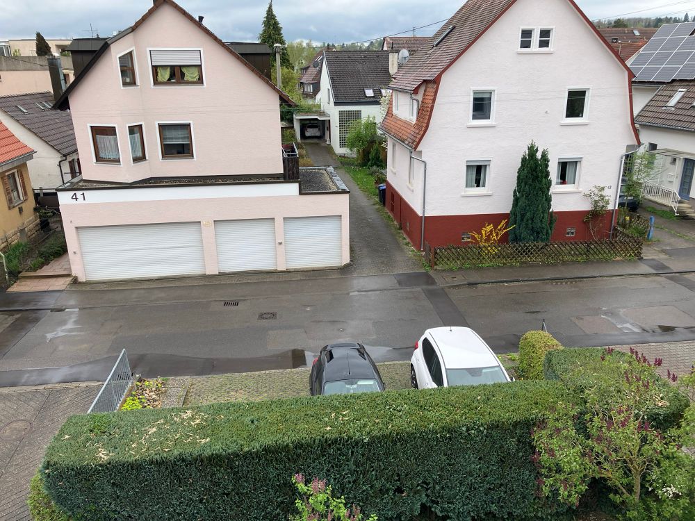 Blick vom Balkon aus auf die Straße. Diese ist nass und es haben sich Pfützen gebildet. Himmel ist eher gräulich u es regnet leicht