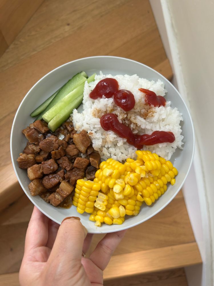 A kid-size plate of rice with a smiley face drawn in ketchup on it, sweetcorn, cucumbers and braised pork belly