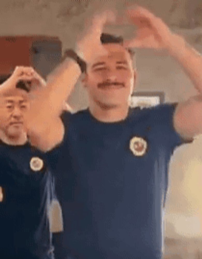 two men are making a heart shape with their hands while wearing blue shirts .