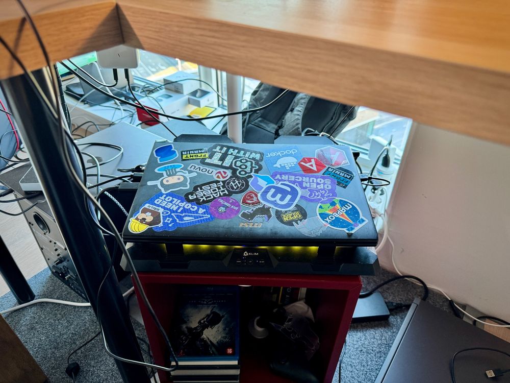 A black laptop full of stickers under a desk. It is on top of a small cube table and a laptop fan. 