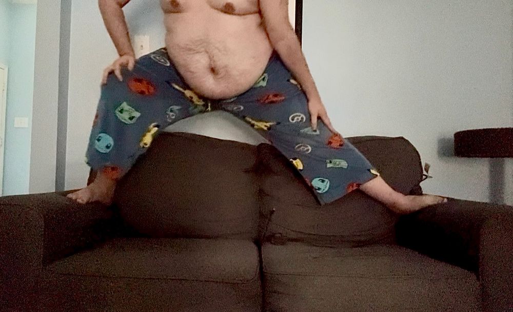 Chubby man wearing only Pokemon pajama pants standing on top of a couch leaning to one side