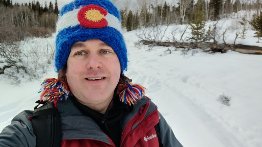 A pale white non binary person wearing a Colorado cap with tasles