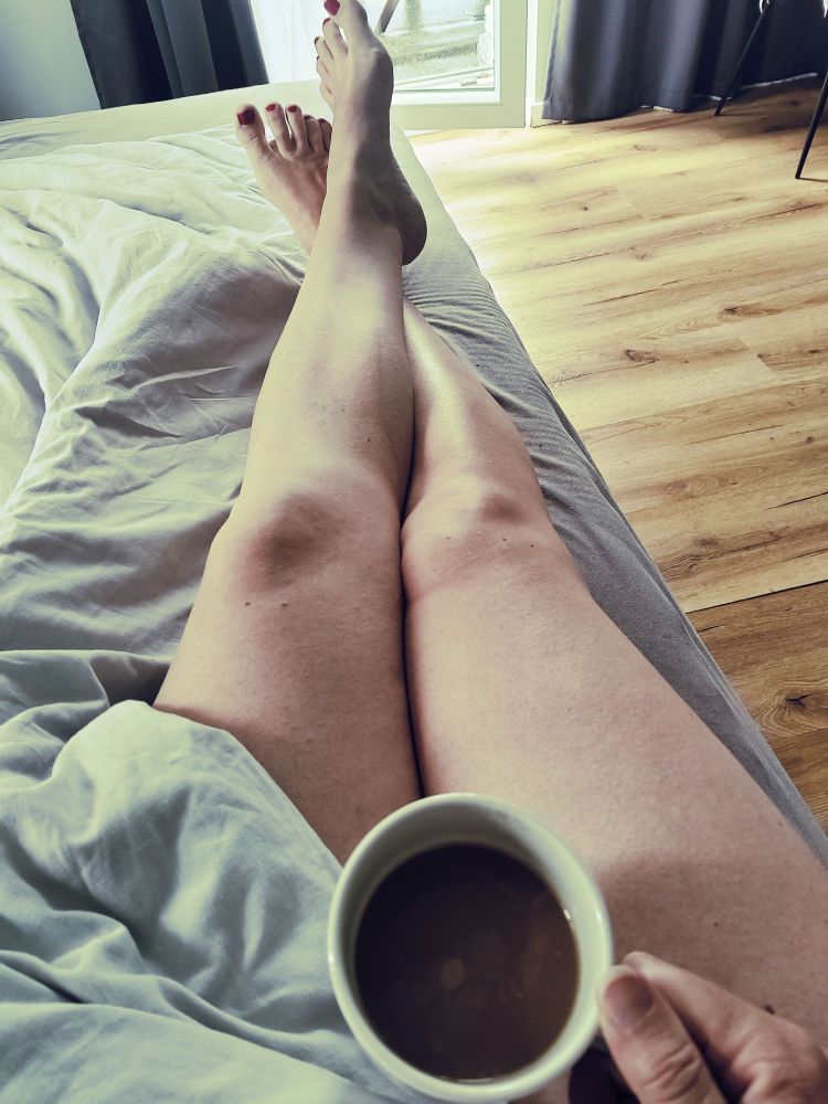 Das Bild zeigt eine entspannte Szene im Schlafzimmer. Im Vordergrund sind Beine zu sehen, die auf einem Bett ausgestreckt und leicht überkreuzt sind. Die Person hält in der rechten Hand eine weiße Tasse mit Kaffee oder einem ähnlichen Heißgetränk. Die Nägel an den Zehen sind rot lackiert. Das Bett ist mit einer weißen Decke bezogen, und im Hintergrund erkennt man einen hellen Holzfußboden, ein Fenster oder eine Glastür sowie dunkelgraue Vorhänge. Durch das Fenster fällt Tageslicht in den Raum, was eine gemütliche und ruhige Stimmung erzeugt.

