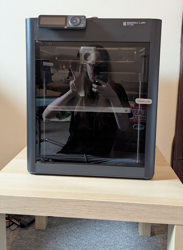 3D printer on a wooden table. The type is a Bambu labs P1S. You can see me in the reflection