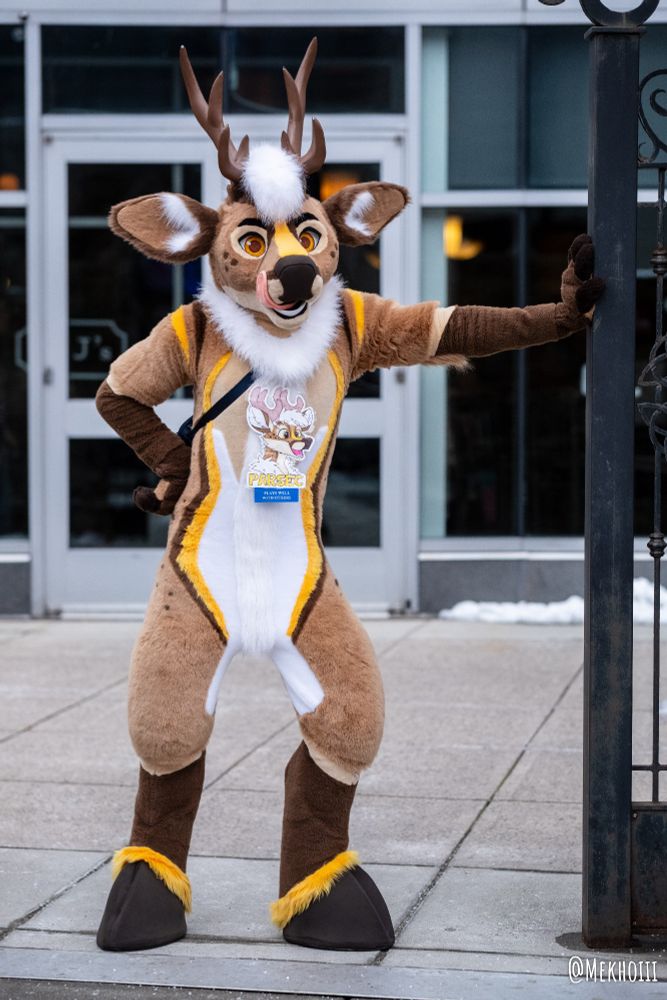 Deer fursuit leaning on a gated fence entryway.